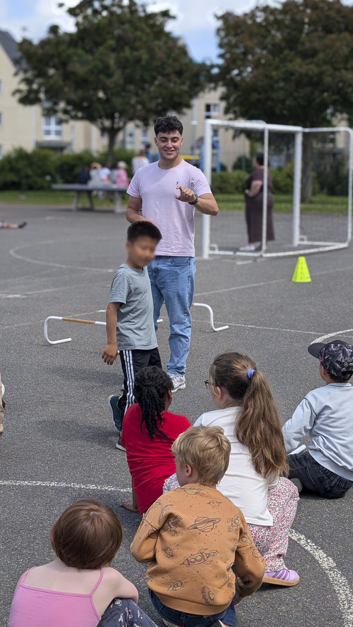 un étudiant de L3 ES dans une cours d'école avec un groupe d'enfant, il leur explique l'activité sportive qu'ils vont effectuer